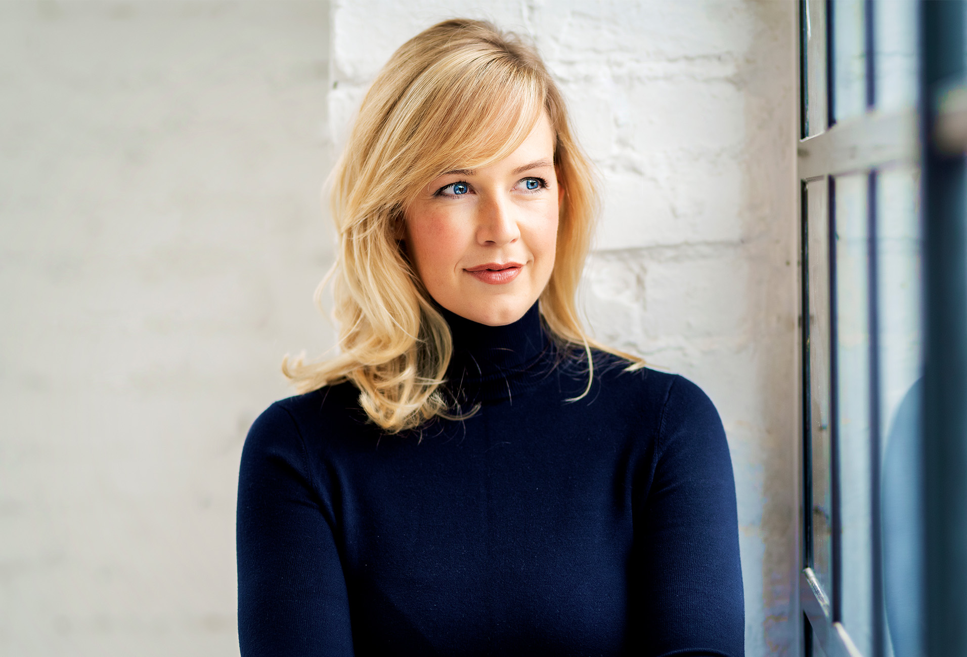 blond woman in blue turtleneck looking out of window with white brick wall